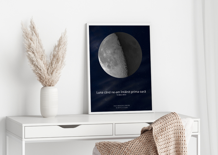 Tablou personalizat cu faza Lunii, asezat pe un birou alb minimalist, alaturi de o vaza cu plante decorative si o patura bej. Tabloul reda Luna exact asa cum a fost vazuta pe 4 aprilie 2015, in Arad, cu textul "Luna cand ne-am intalnit prima oara". Ideal pentru cadouri romantice si decor cu semnificatie personala.