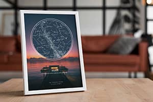 Tablou personalizat cu harta cerului, sprijinit pe o masa din lemn, intr-un living modern cu canapea maro si decor industrial. Posterul reda constelatiile vizibile deasupra unui apus spectaculos, alaturi de mesajul „The night we met”. Ideal ca decor de cuplu sau cadou pentru aniversari si momente speciale.